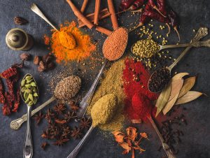 Variety of herbs and spices on a dark grey background. Cinnamon stick, rosemary, black pepper, paprika, thyme, etc. are placed on spoons.