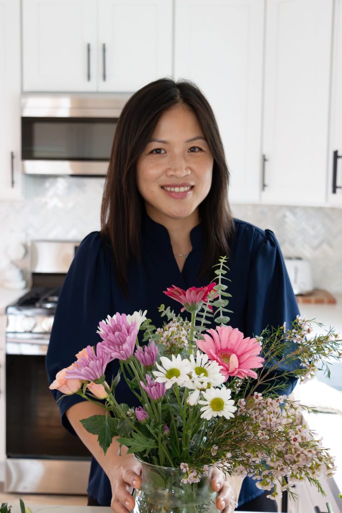 Asian woman wearing dark blue top holding vase of pink and white wildflowers in a modern kitchen
